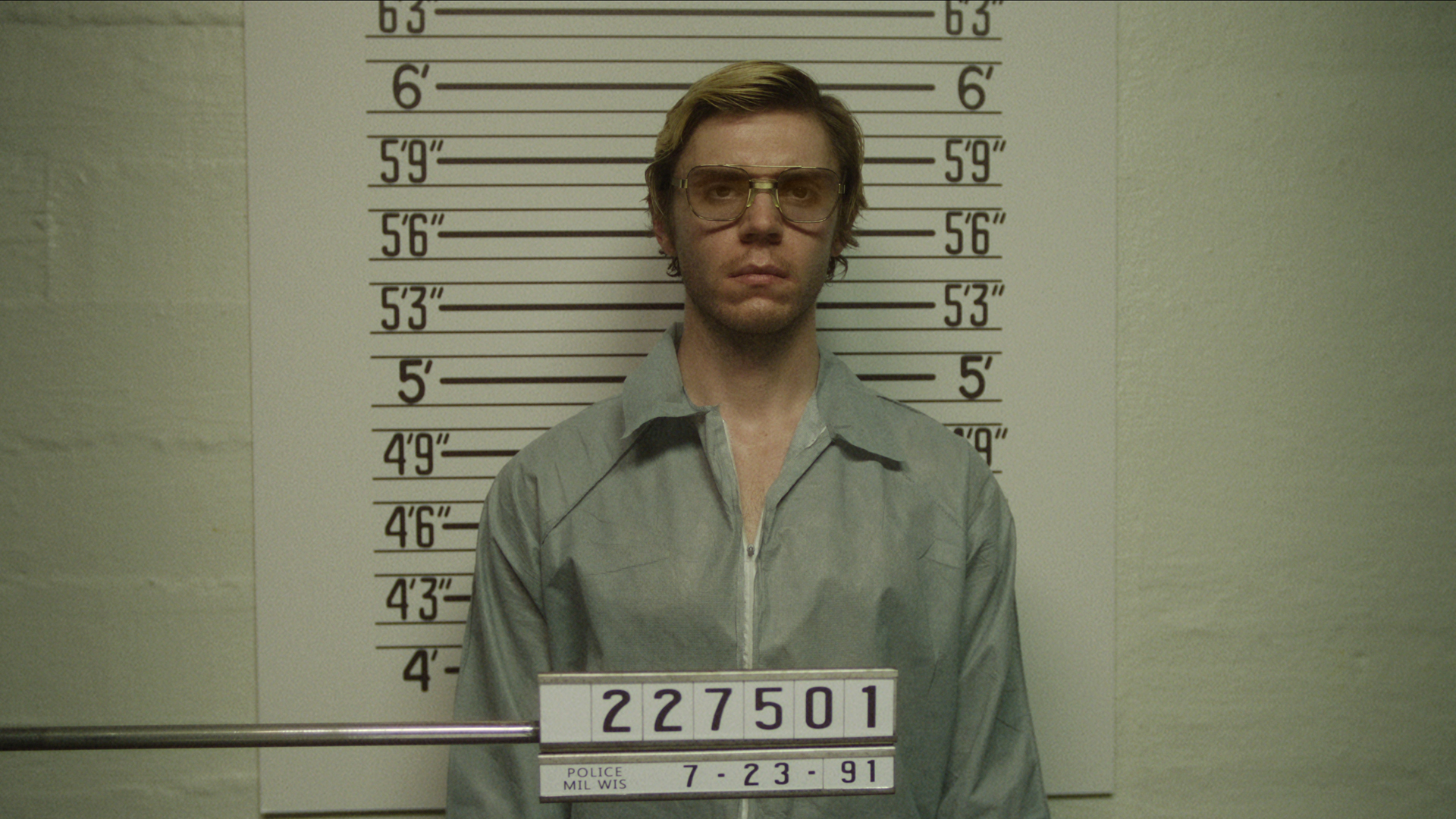 Evan Peters' Jeffrey Dahmer stands in front of a measuring wall as his police photograph is taken in Monster: The Jeffrey Dahmer Story