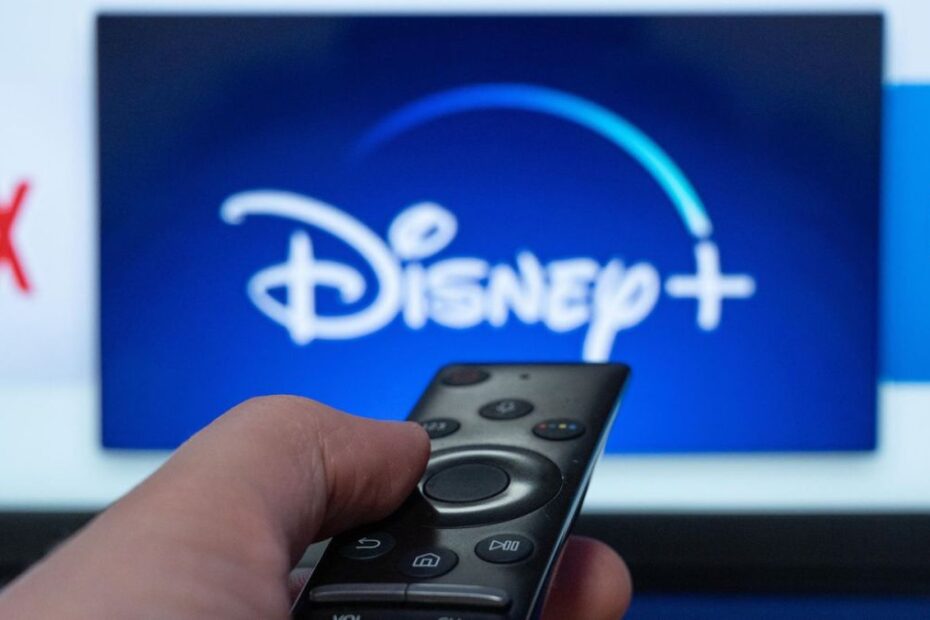 A person points a remote at their TV on which the Disney Plus logo is displayed